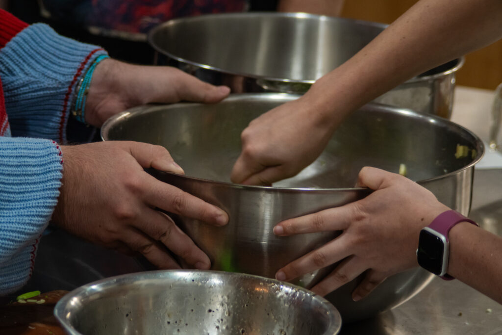 Two people holding a big metal mixing bowl. One is holding and the other person is mixing what is in the bowl.
